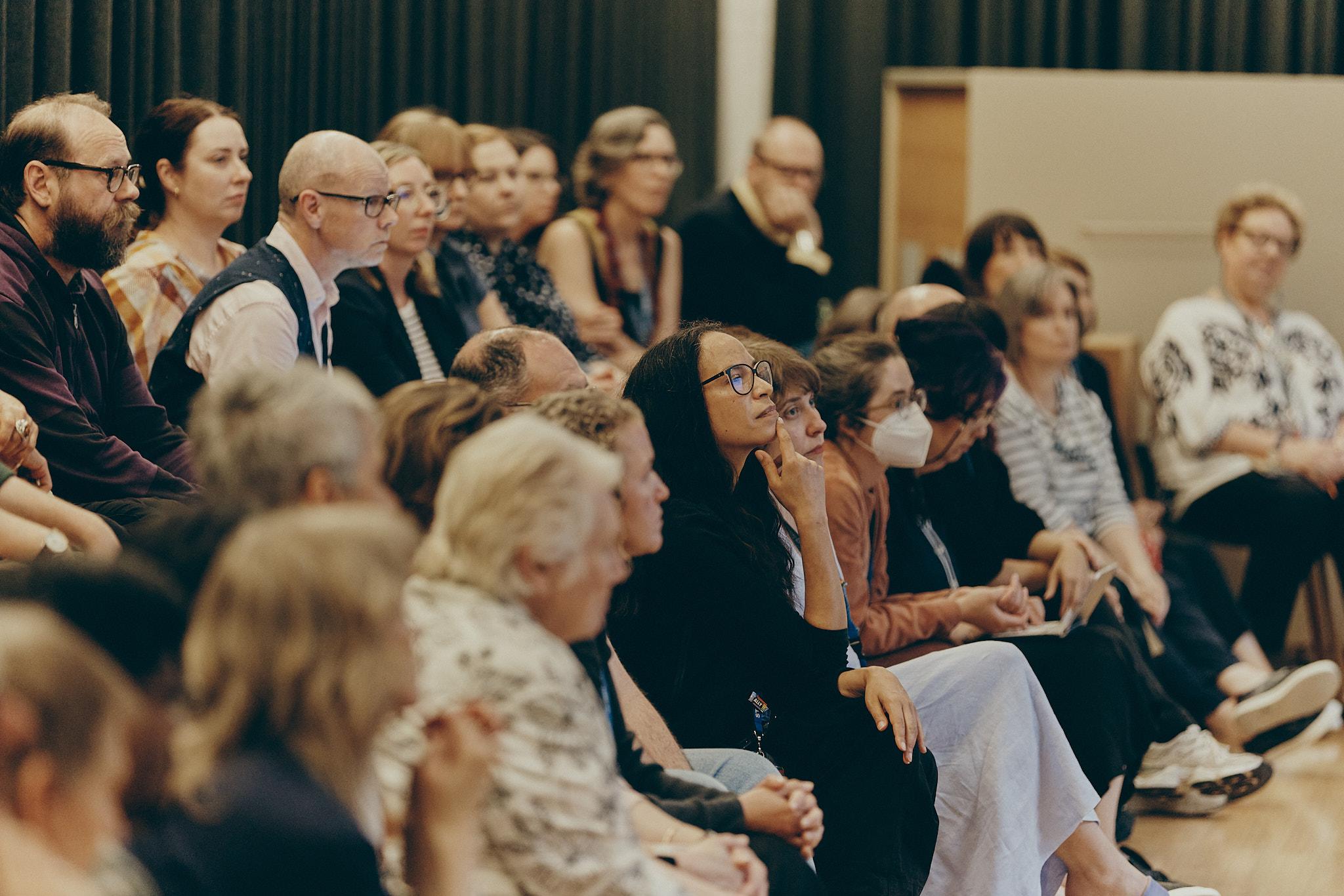 A crowd sits on wooden bleachers, looking, with thoughtful expressions, to the right of frame