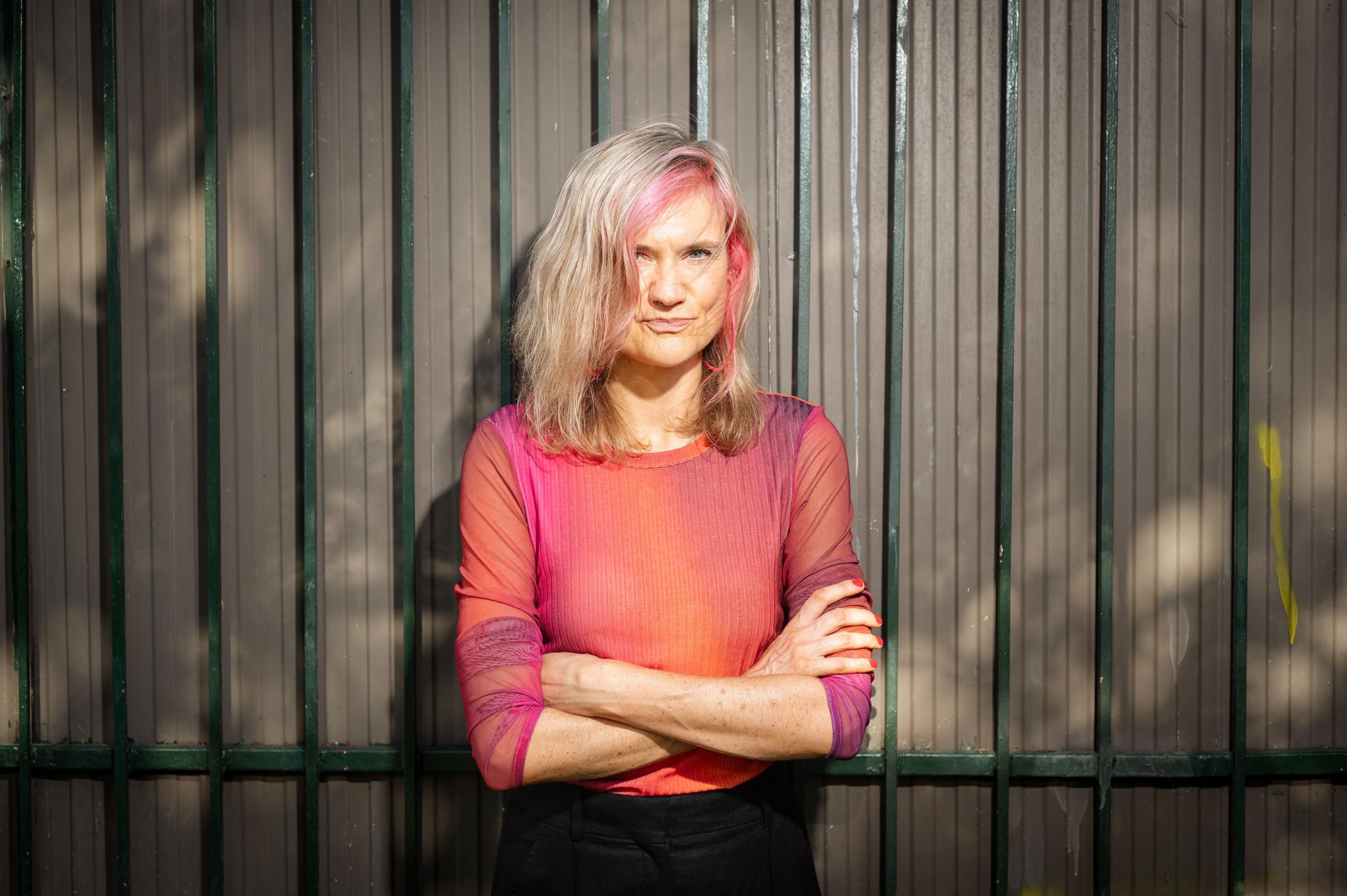 A mid shot of a woman with long blonde hair with a purple streak wearing a orange-purple top with crossed arms