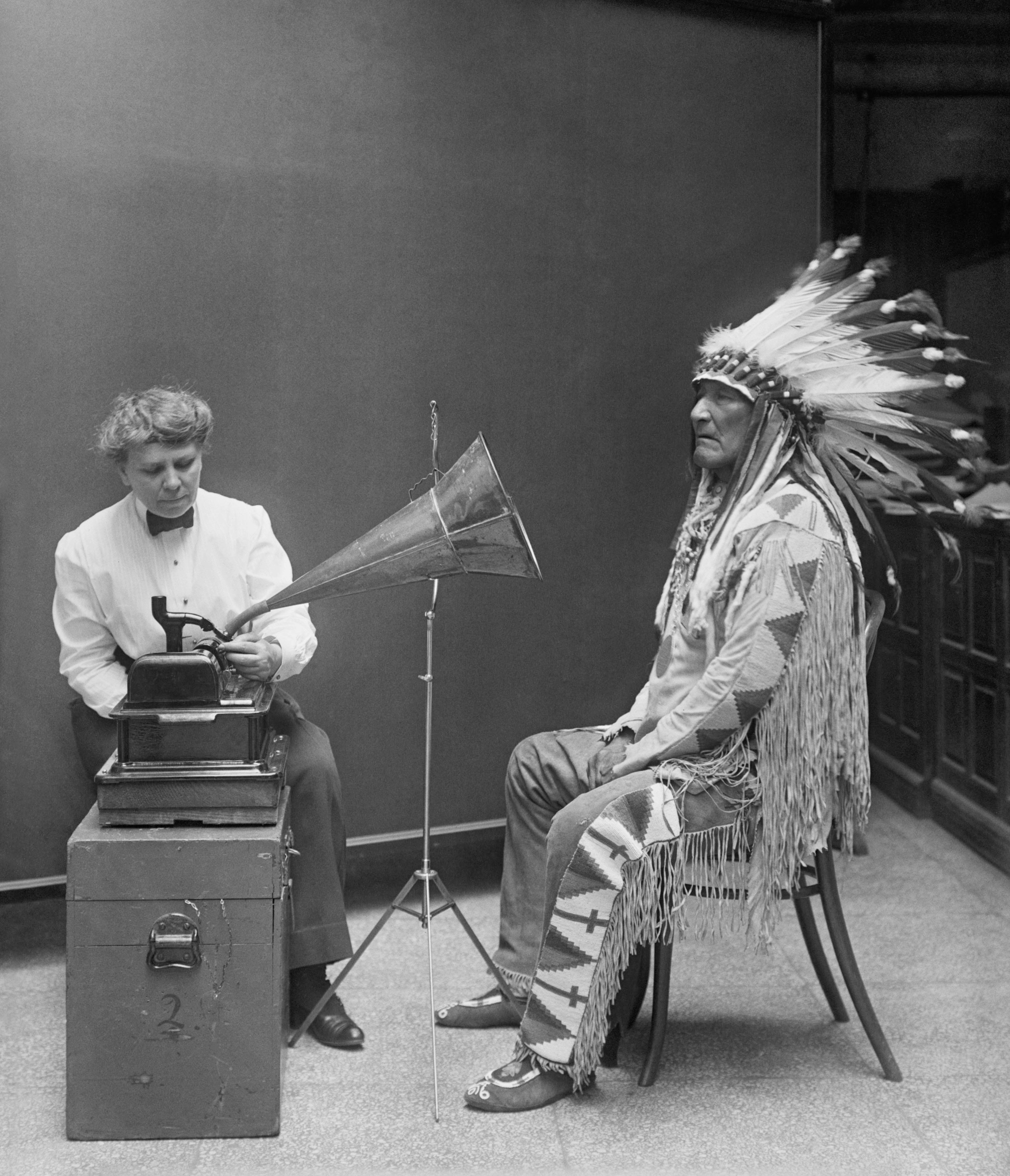 A 19th century black and white photo showing a native American chieftain with a large ornate headdress (right) seated opposite a female western researcher (left) who is operating an early audio recording device.