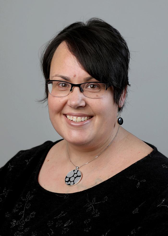 A portrait of a smiling woman with short black hair and glasses. She has black earrings and a silver necklace