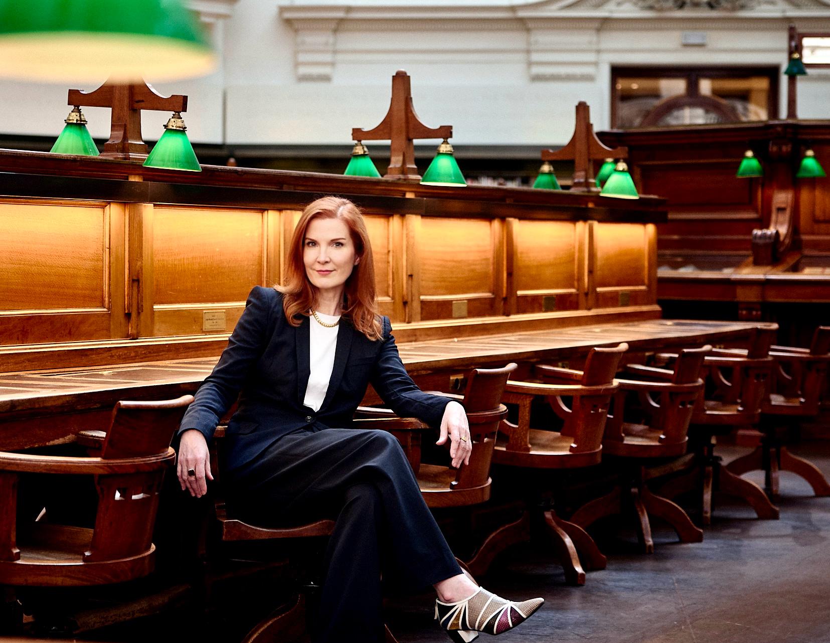 A woman (Kate Crawford) with long red hair wearing a suit seated in the La Trobe Reading Room at State Library Victoria