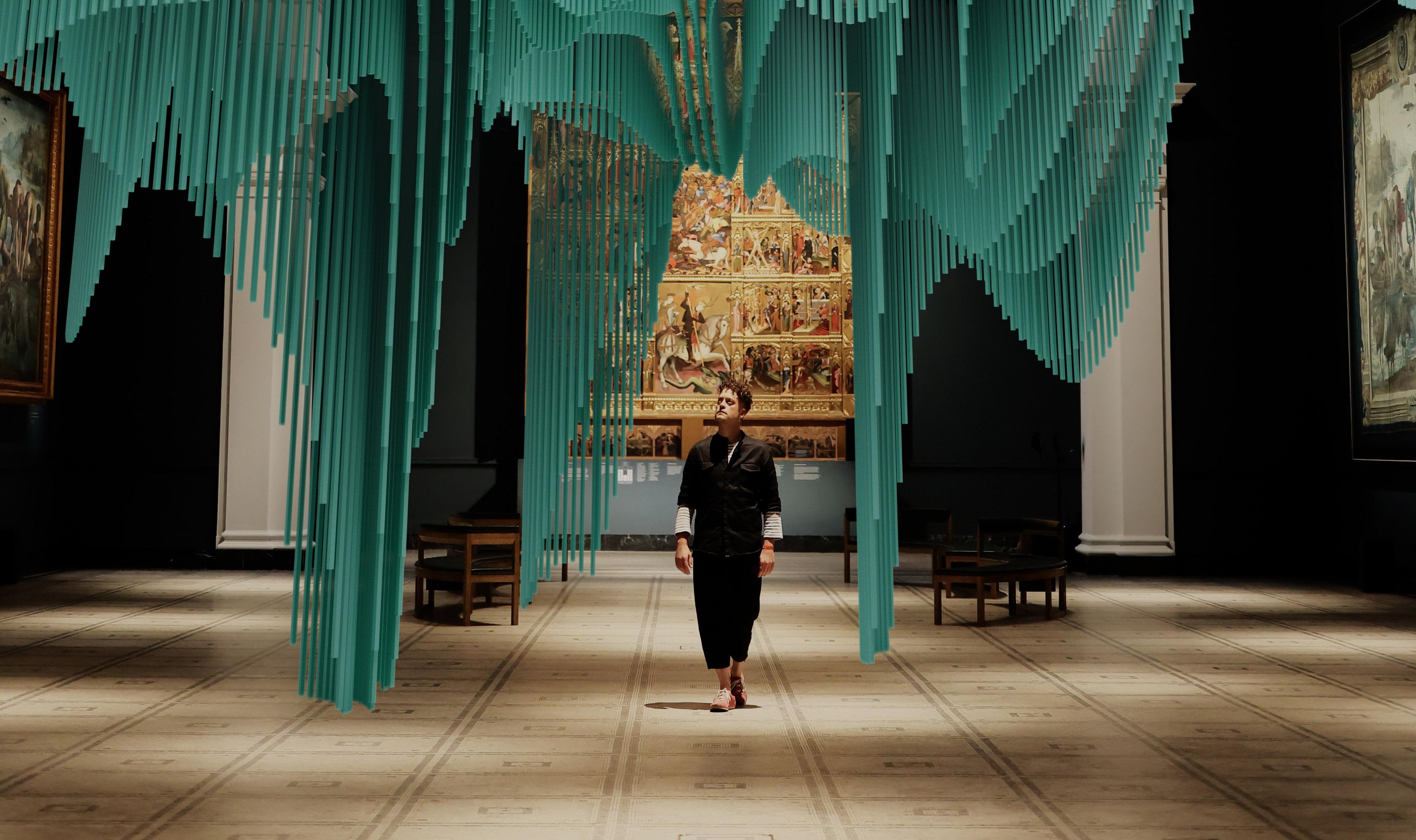 A cropped version of a person (Yoyo Munk) in black in centre of frame inside a gallery space looking up to the left at a virtual reality columnar blue artchitecture
