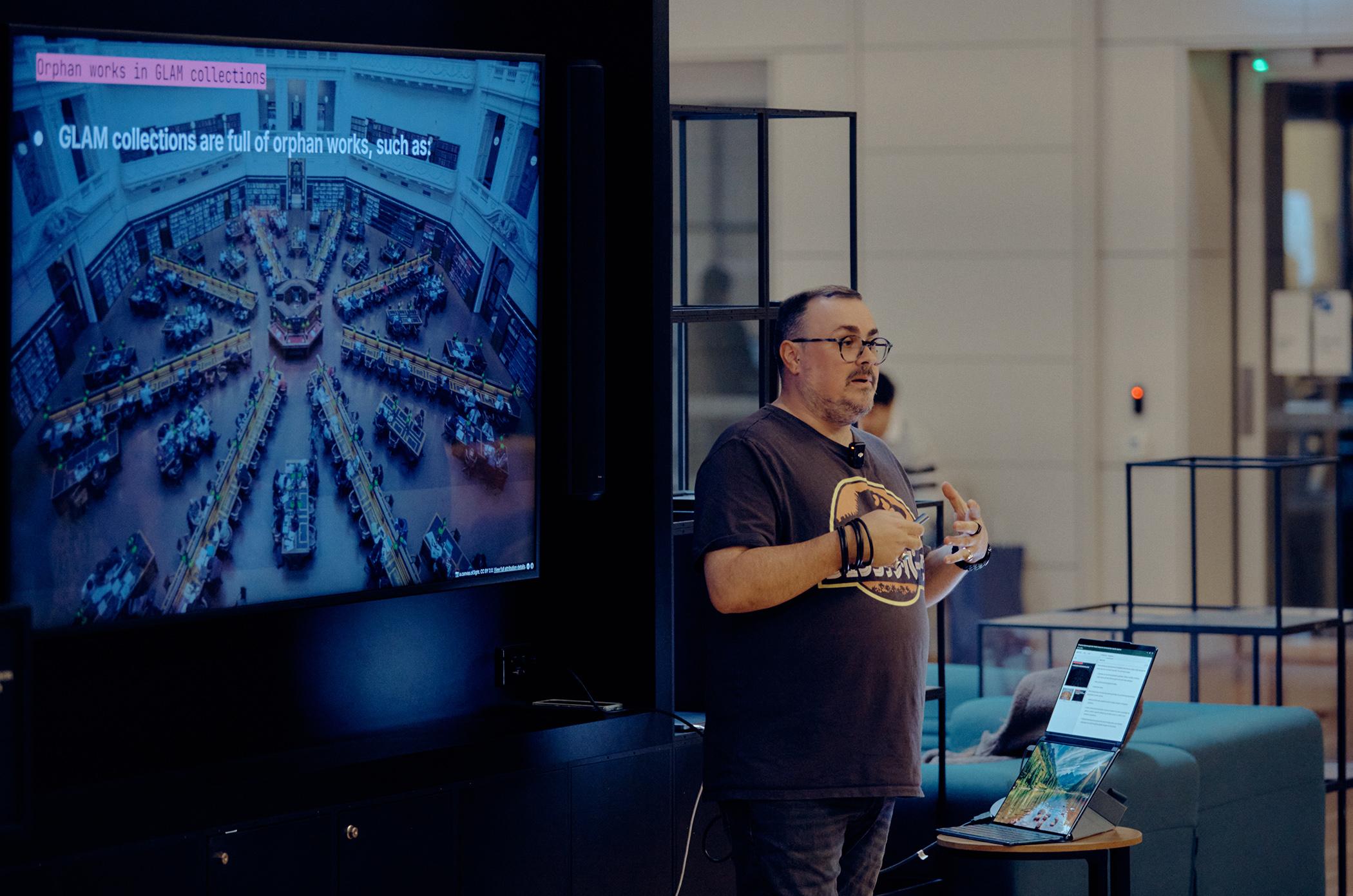 A man with short hair and glasses presenting a lecture to a crowd to the right of frame