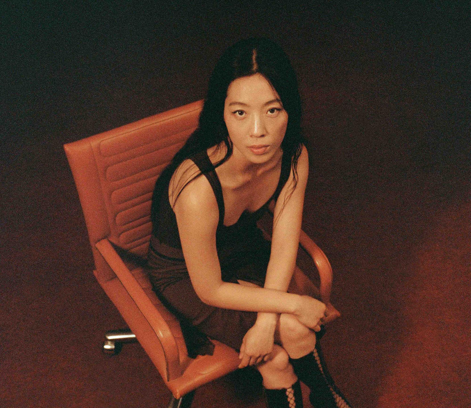  A high angle photo of a woman of asian descent with long black hair wearing a black top sitting on a stylish brown leather chair.
