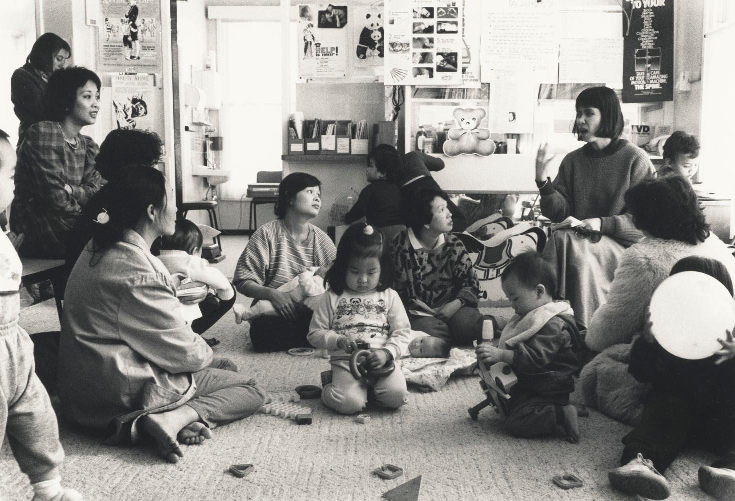 A black and white photo of a group of women and children of indo-Asian origin sitting on the floor in a classroom setting and listening to another woman speak.