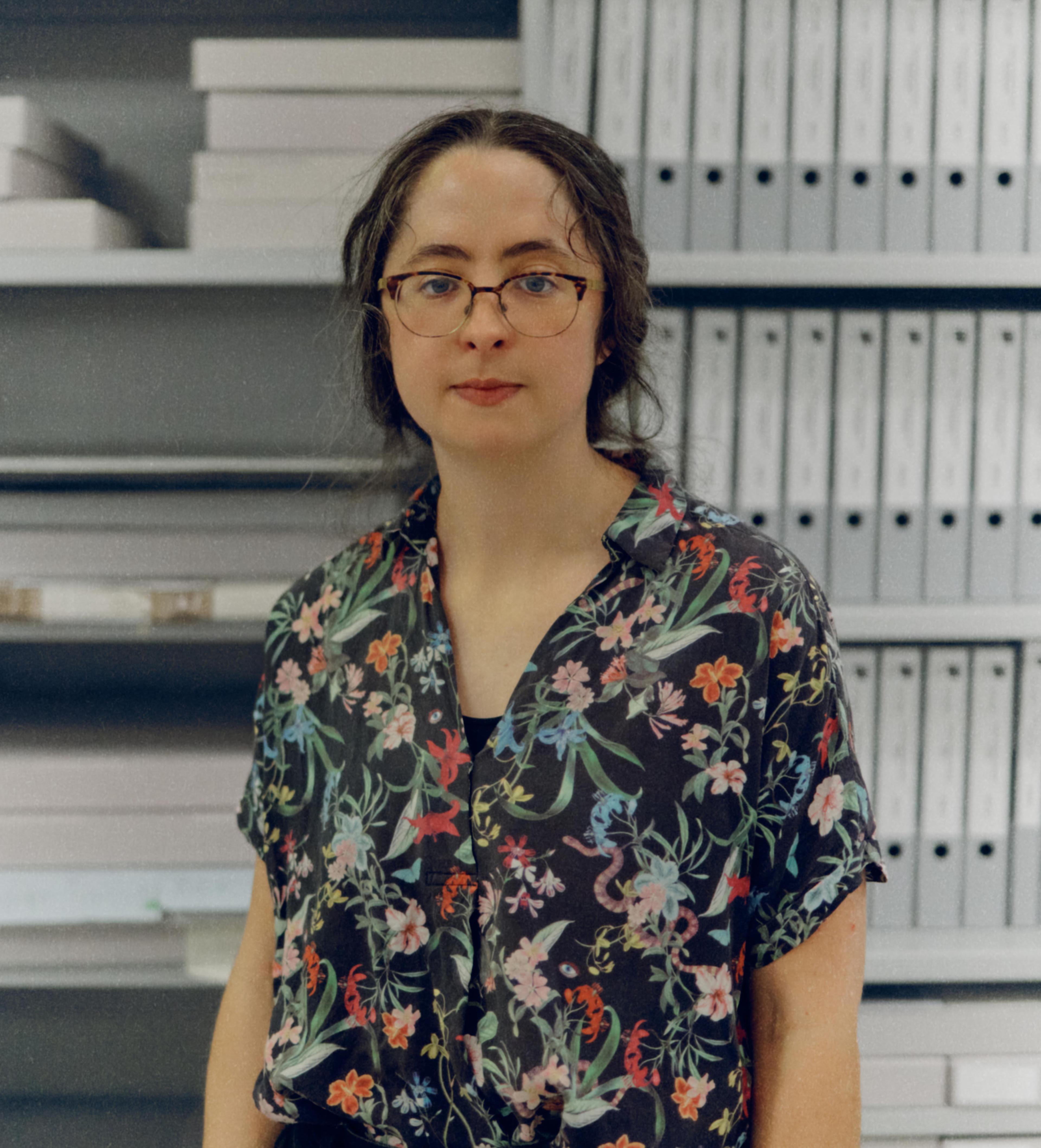 Headshot of a bespectacled woman with dark hair, wearing a floral collared shirt standing in front of shelves with archival document folders