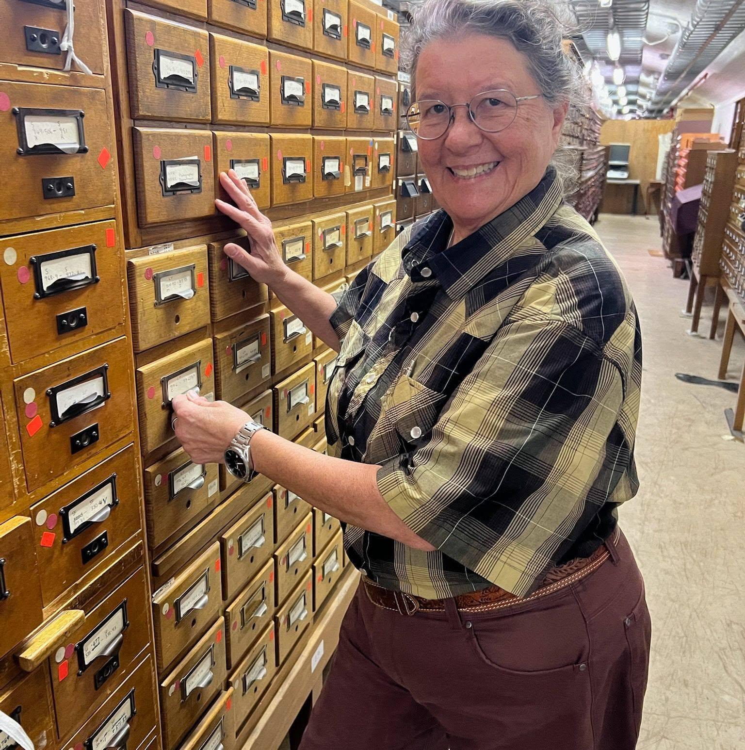 A person (Cynde Moya) wearing glasses in a chequered shirt standing by archival drawers