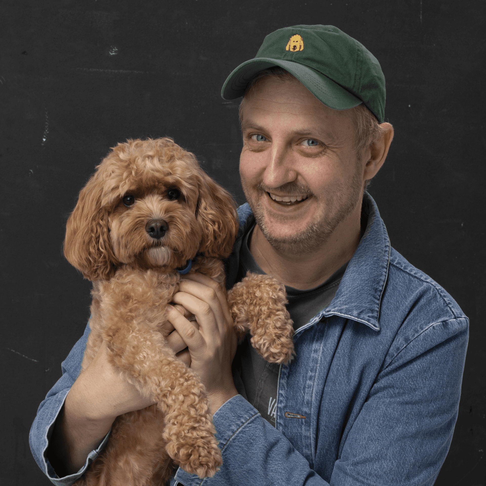 Headshot of a smiling man in a green hat and jean shirt, holding a light brown poodle.