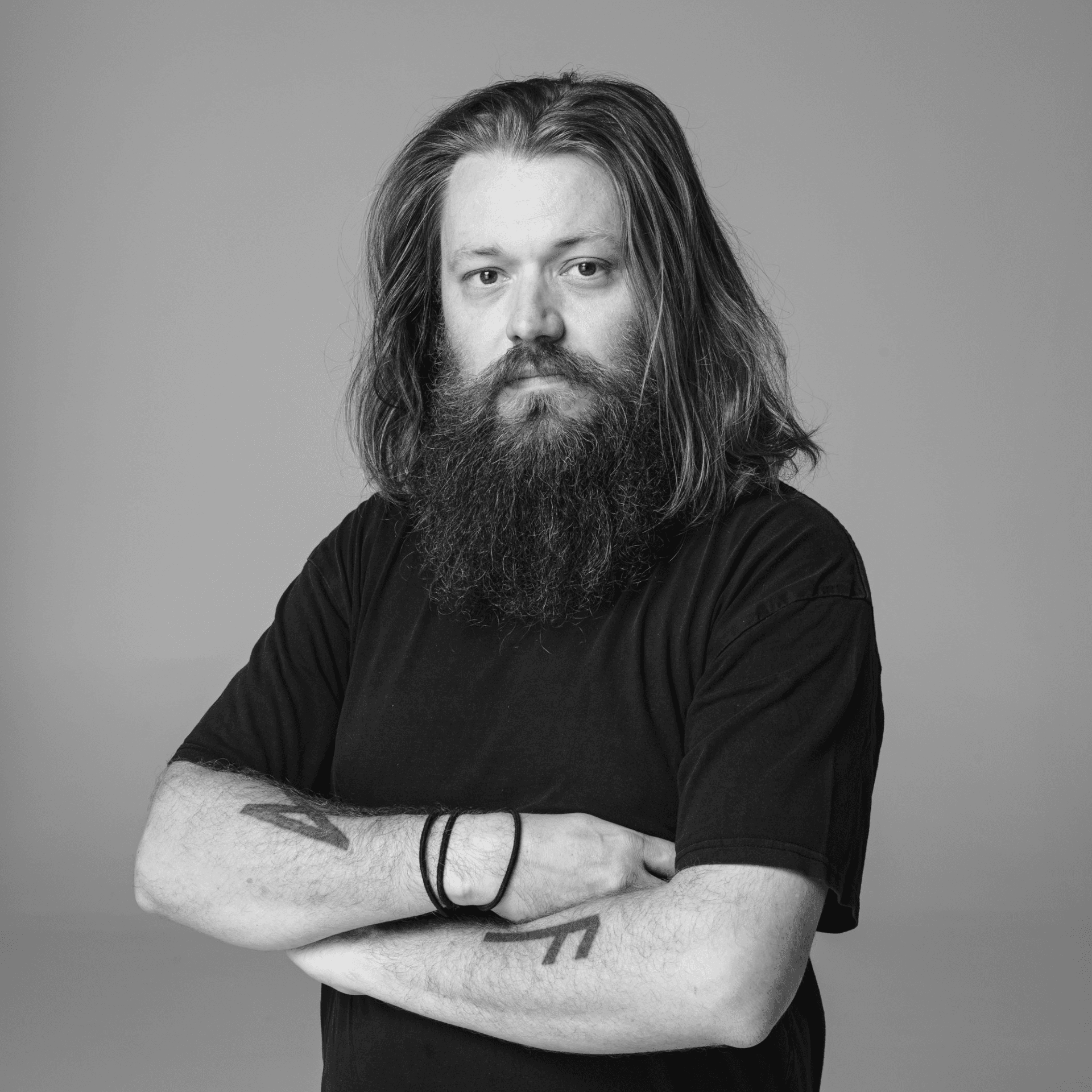 Headshot of a bearded man with long brown hair, standing with his arms crossed in a black t-shirt.