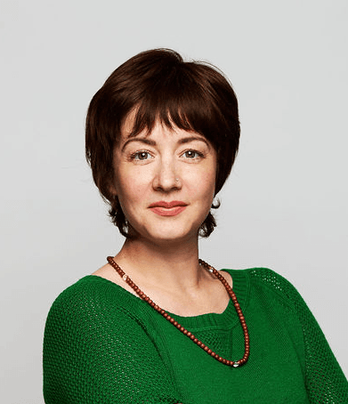 A woman with dark brown mid length hair looks towards the camera. She wears a forest green jumper and a beaded necklace.