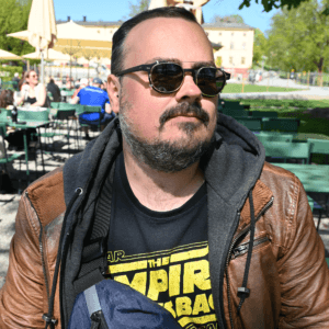 Headshot of a bearded man, wearing sunglasses, a dark t-short and tan leather jacket. 