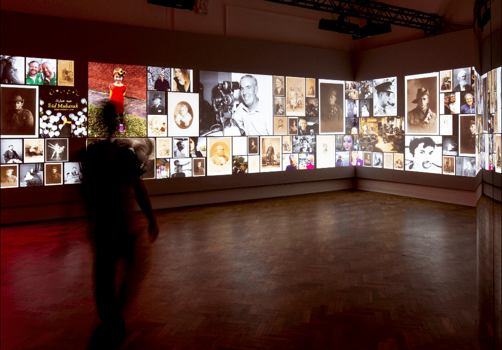 A person walks into a darkened gallery. Large scale projections cover the walls, displaying colourful photos of library collection items.