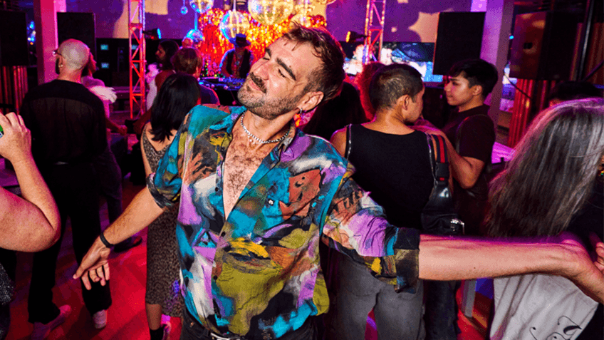 A man in a colourful patterned short dances in the middle of a busy, vibrant scene.