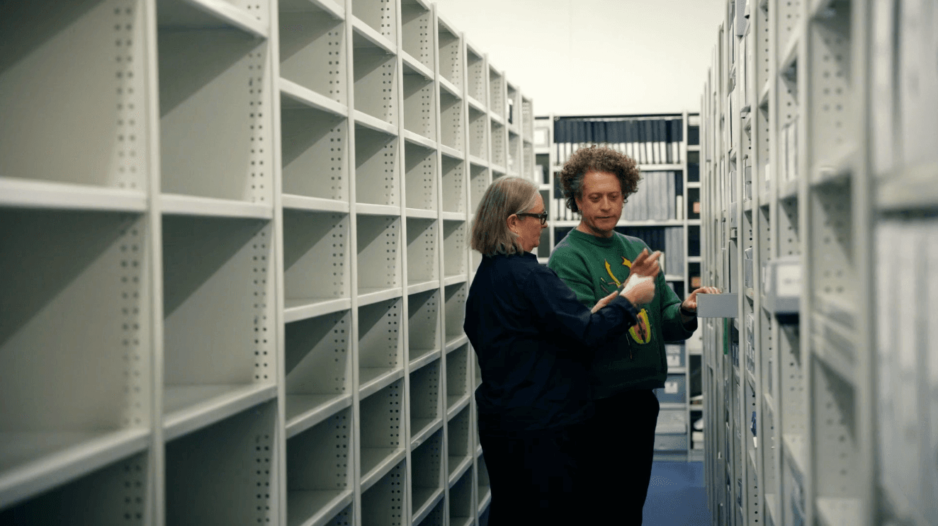 A woman and a man standing between a long row of grey shelving talking to each other