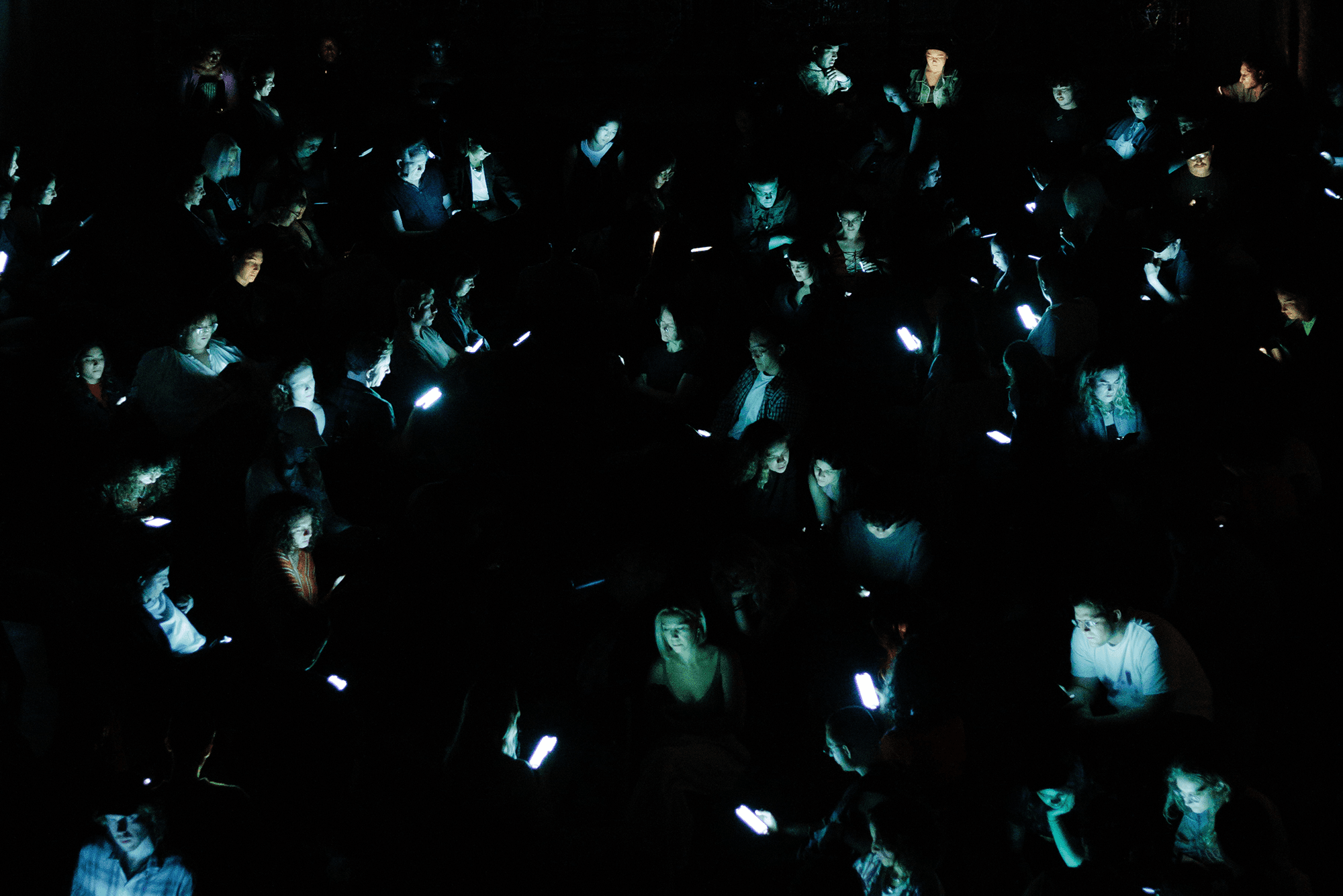 A crowd of people in the dark with their faces illuminated by phone screens